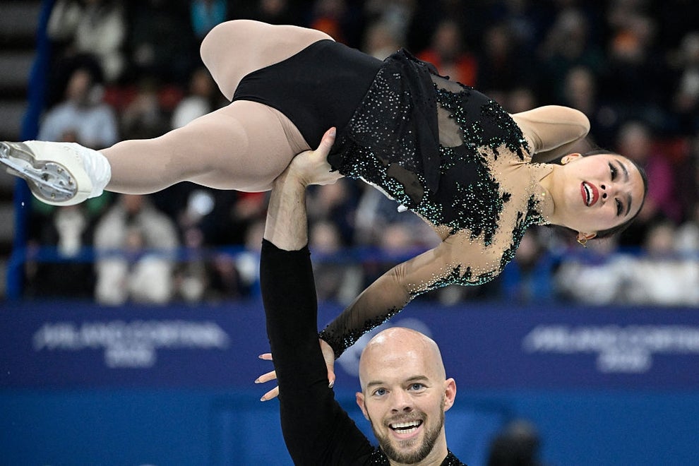 Figure skaters performing a lift; woman in black costume held aloft by bald partner during routine
