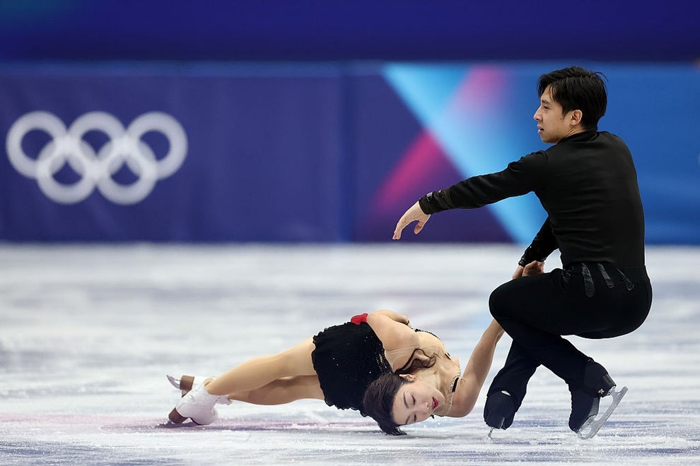 Two figure skaters perform on ice; one skater is elegantly leaned back, while the other supports them, both wearing sleek black costumes