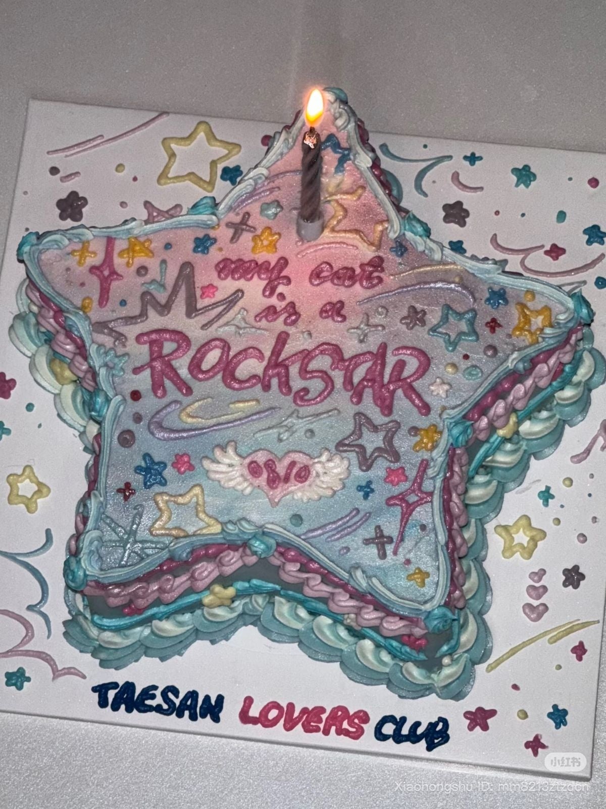 Pastel en forma de estrella con una vela encendida, decorado con texto que dice "My Dad is a Rockstar" y "Taesan Lovers Club"