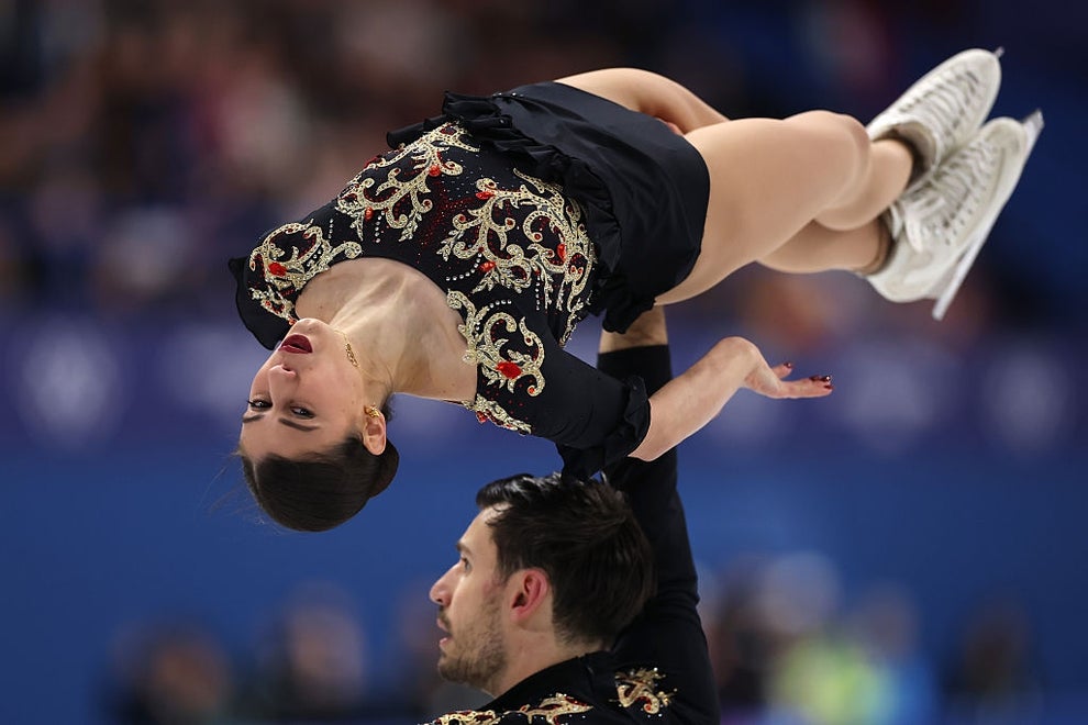 Figure skaters perform a dramatic lift on the ice, wearing ornate costumes with intricate patterns