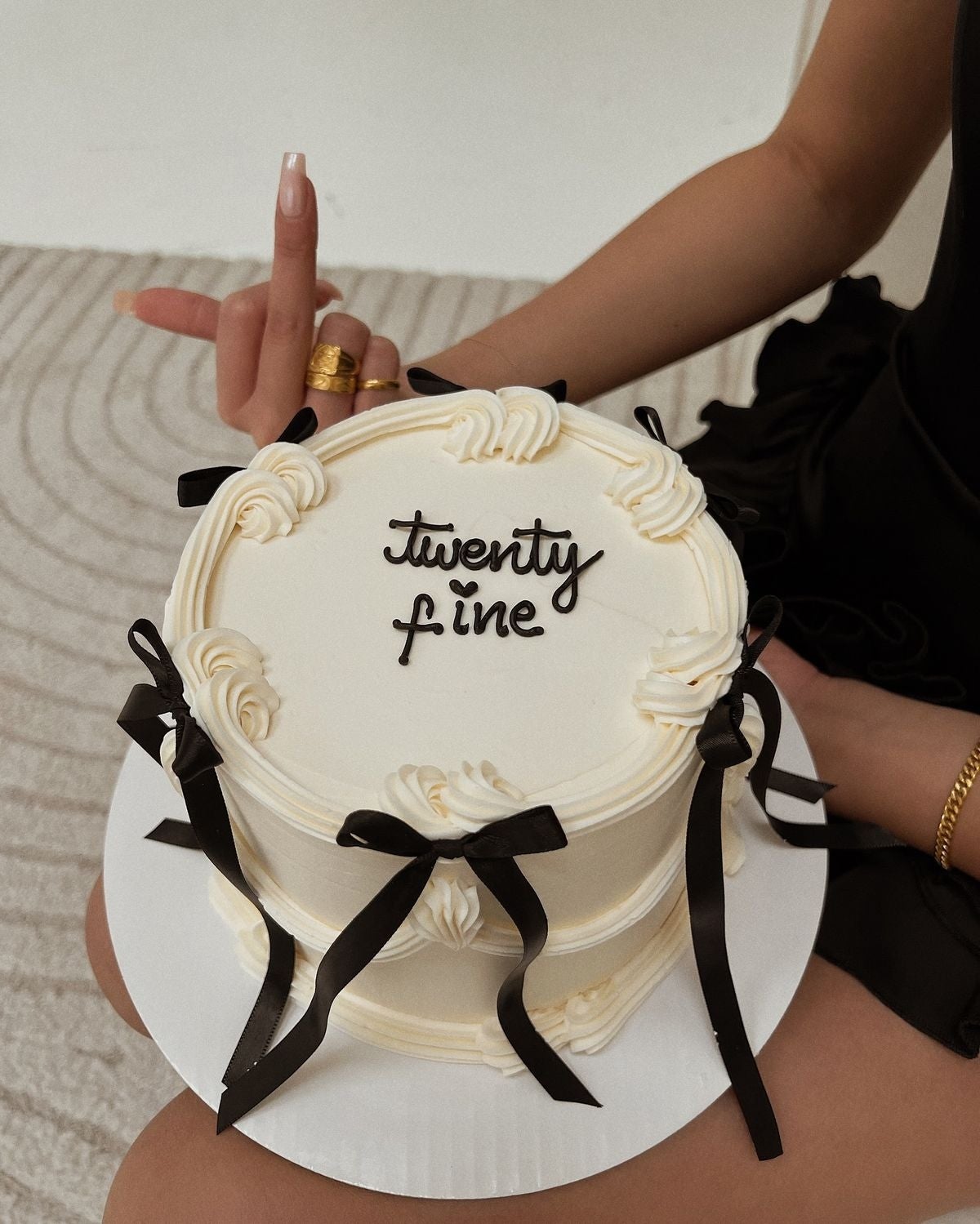 Persona mostrando un pastel con texto "twenty fine" decorado con lazos negros. Está levantando un dedo y lleva un vestido elegante