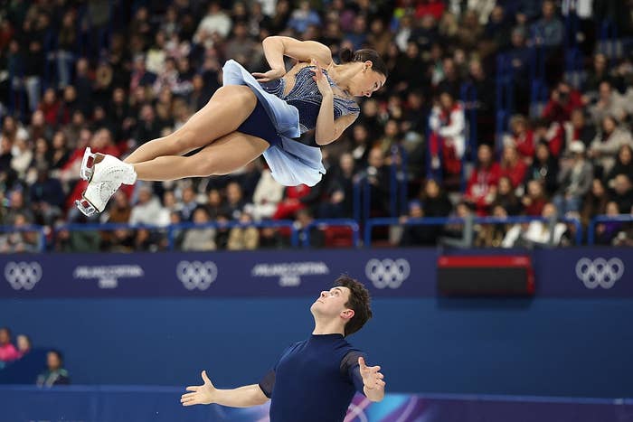 Figure skaters perform a dramatic lift during a competition, with the female skater soaring gracefully above her partner