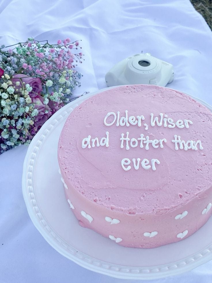 Tarta rosada con texto "Older, Wiser, and Hotter than ever" al lado de flores y una cámara instantánea sobre una tela
