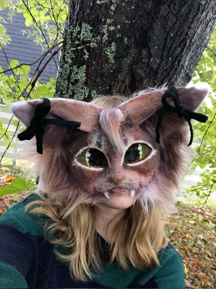 Persona con máscara de gato de fantasía, con orejas grandes y colmillos, posando al aire libre frente a un árbol