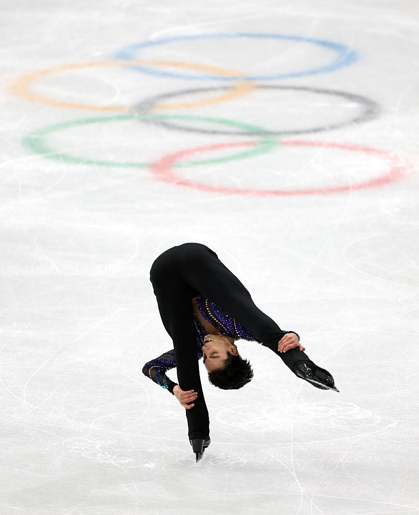 Figure skater performing a back-bending spin on ice, Olympic rings visible in the background