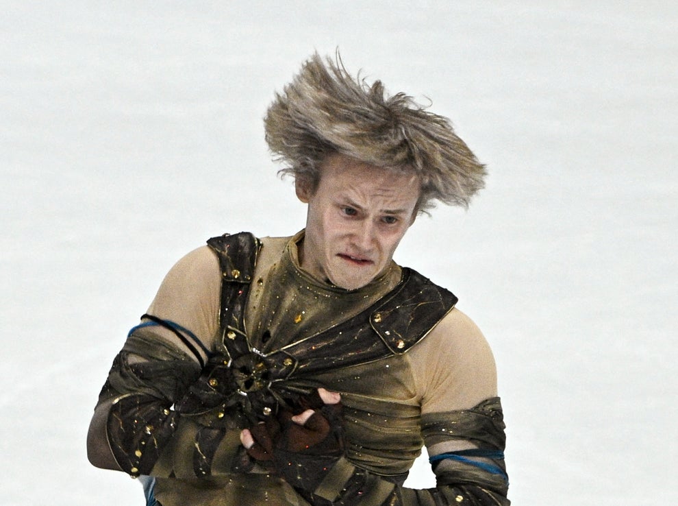 Figure skater mid-jump wearing a themed costume with arm cuffs and a belt, performing on the ice
