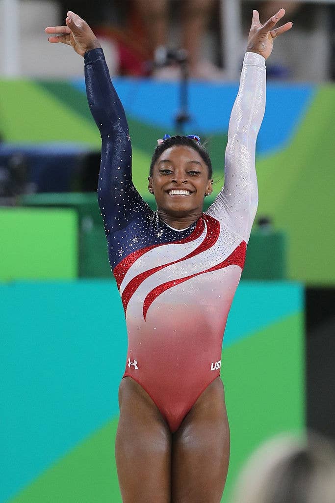 Gymnast performing a routine, wearing a star-spangled leotard with a question    design. Arms raised successful  a triumphant pose, smiling astatine  the audience