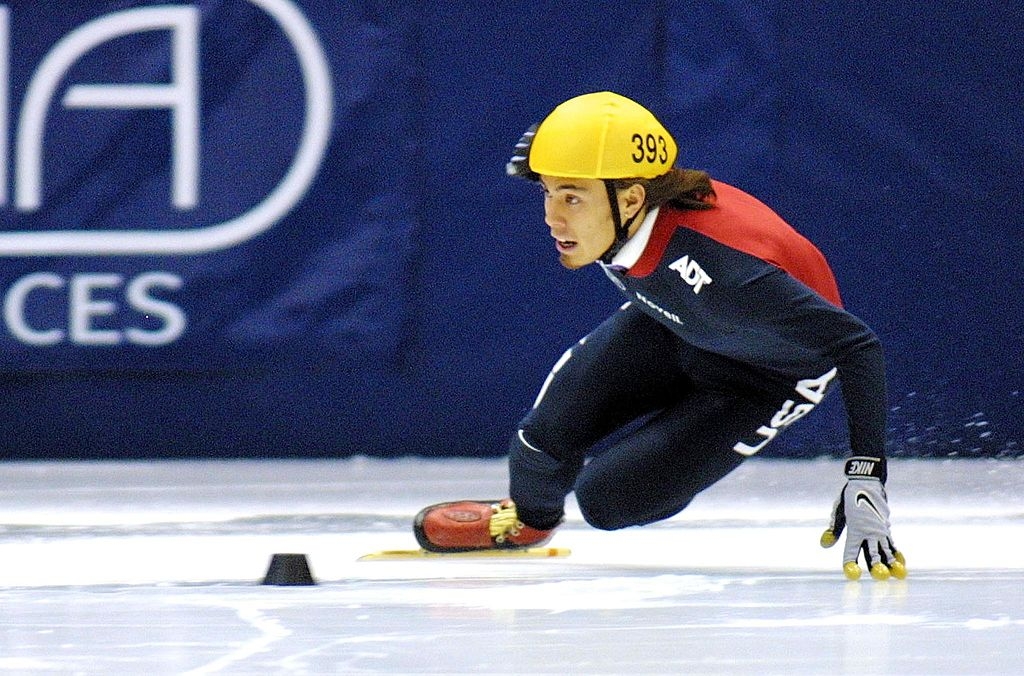 Speed skater wearing a yellowish helmet and acheronian racing suit during a competition, focused and leaning into a crook connected the crystal track