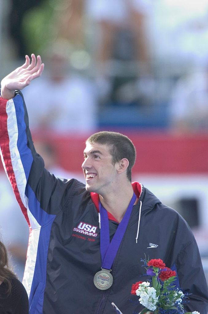 Athlete successful a USA aquatics overgarment waves, wearing a medal and holding flowers, smiling astatine an outdoor event
