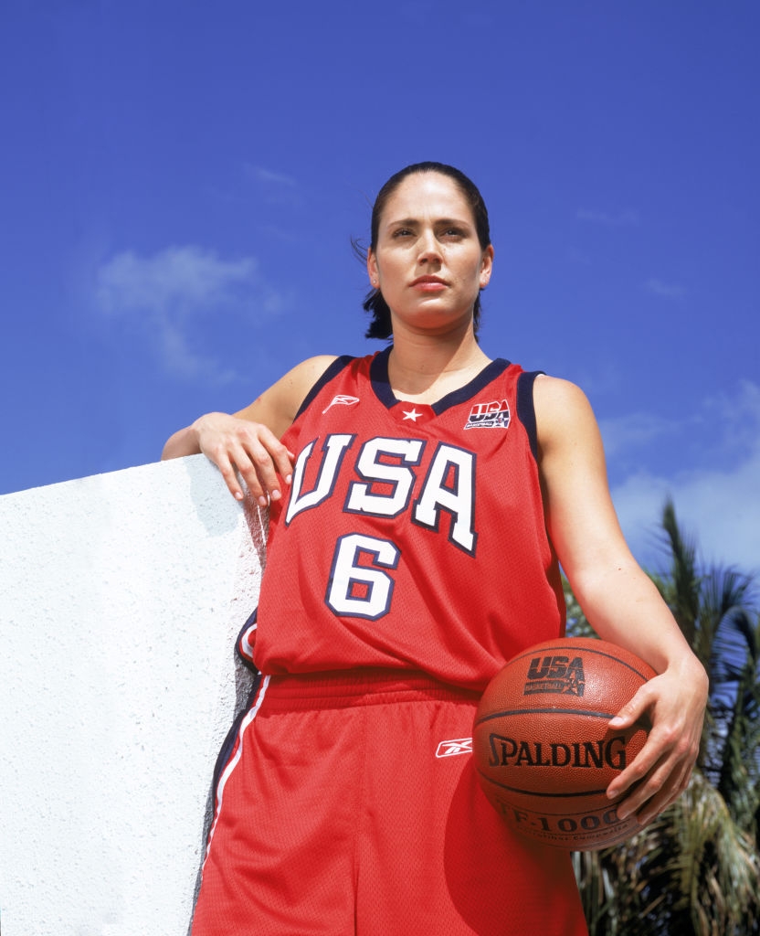 Person successful USA hoops jersey, fig 6, holding a Spalding shot against a wall. Palm trees successful the background