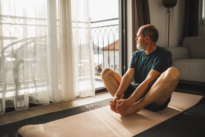 A antheral   sits cross-legged connected  a yoga mat by a window, gazing outside