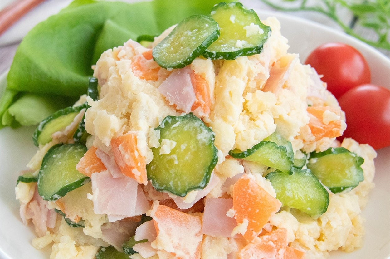 クリーミーなポテトサラダがプレートに盛られ、きゅうりとハムがトッピングされています。隣にプチトマトが添えられています。