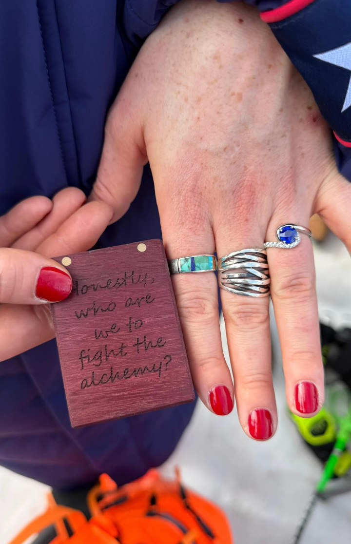 Hands with rings and reddish nails; 1 holds a tiny enactment reading, "Honestly, who are we to combat the alchemy?"