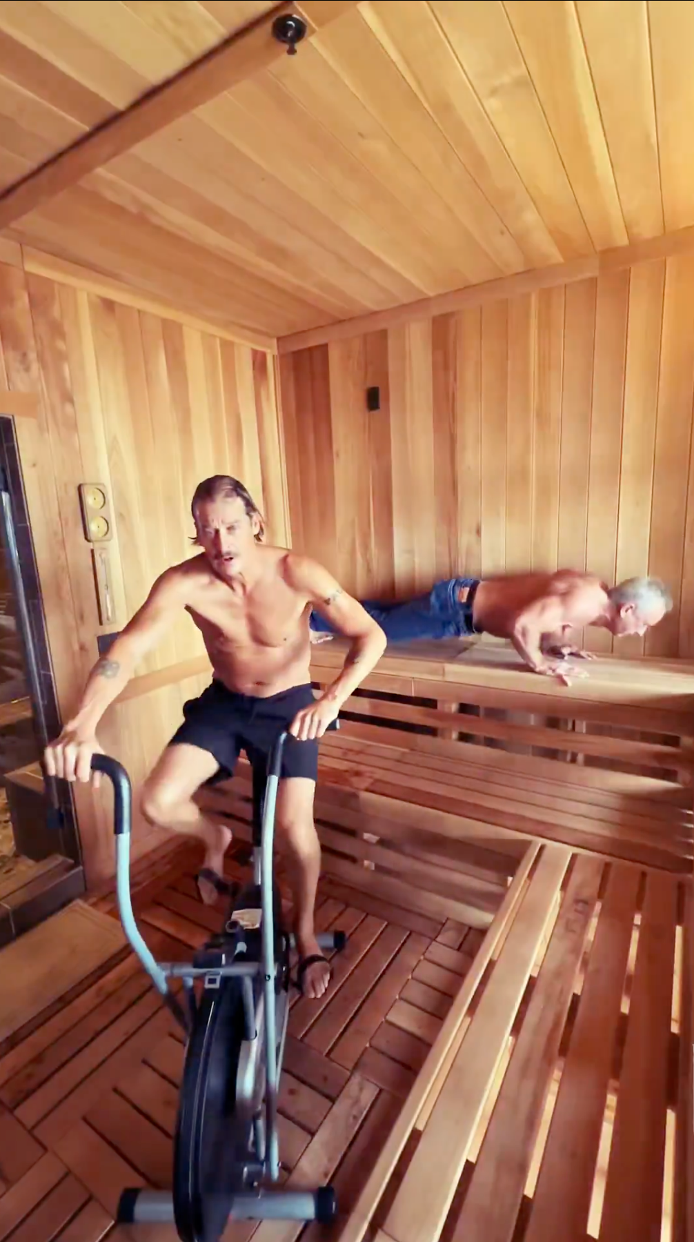 To menn uten skjorte i en trebadstue, den ene sykler på en stillestående sykkel og den andre gjør push-ups på en benk