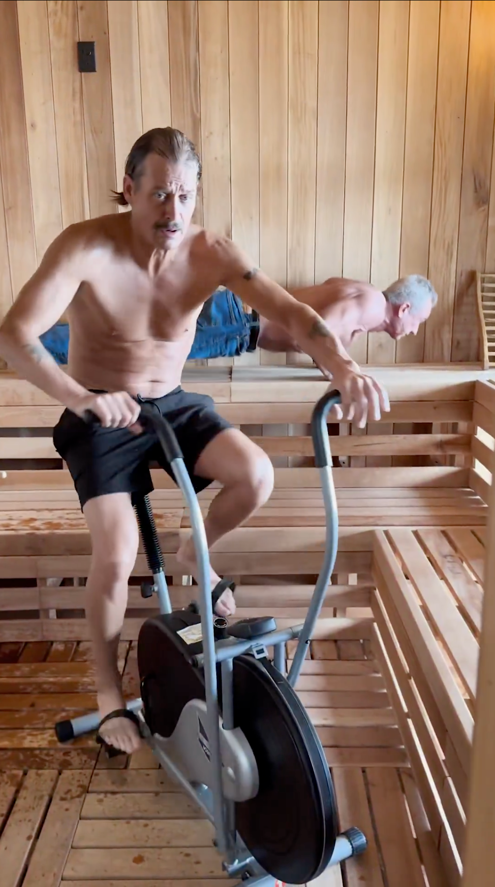 To menn i en badstue; den ene sykler på en stasjonær sykkel uten overkropp, iført svarte shorts og sandaler, den andre lener seg på en benk
