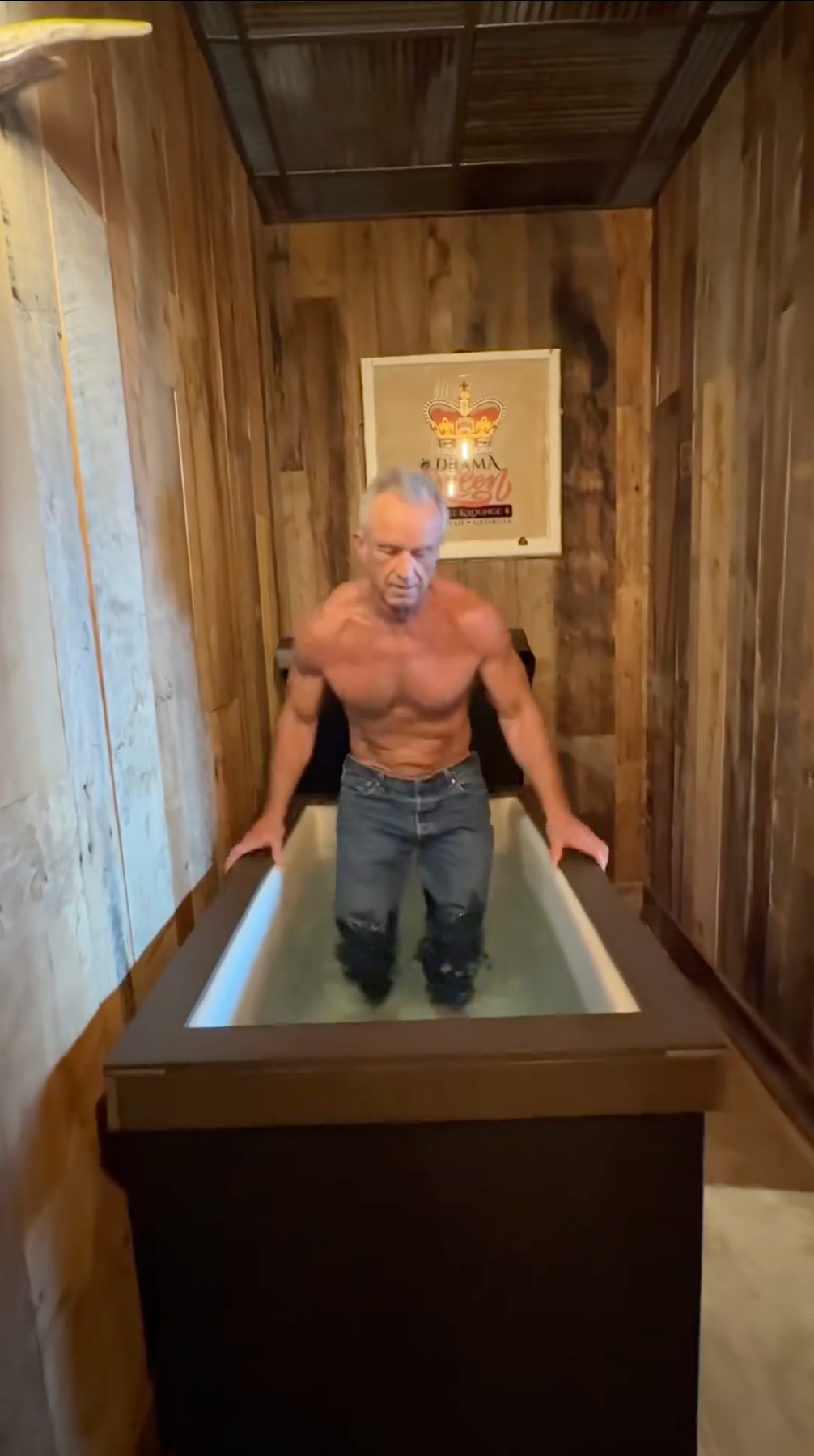 En mann i jeans befinner seg i et trebadekar fylt med vann, i et rustikk rom. En veggplakat er delvis synlig