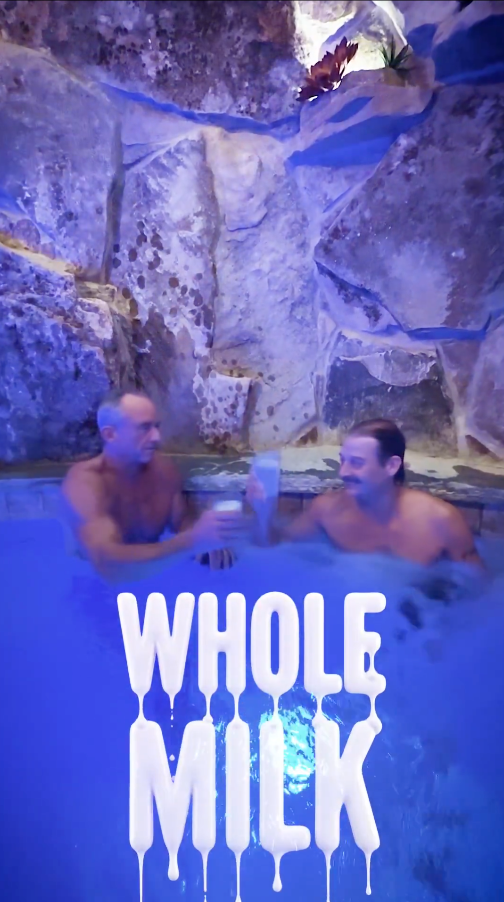 To personer i en skummende badestamp, ler og skåler med drinker. Stor tekst lyder "HELMELK" i et dryppende skriftoverlegg