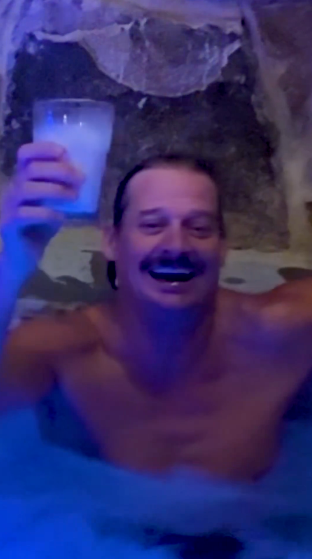 Person som smiler i en badestamp, holder en drink i den ene hånden, virker munter og avslappet. Steinmur i bakgrunnen