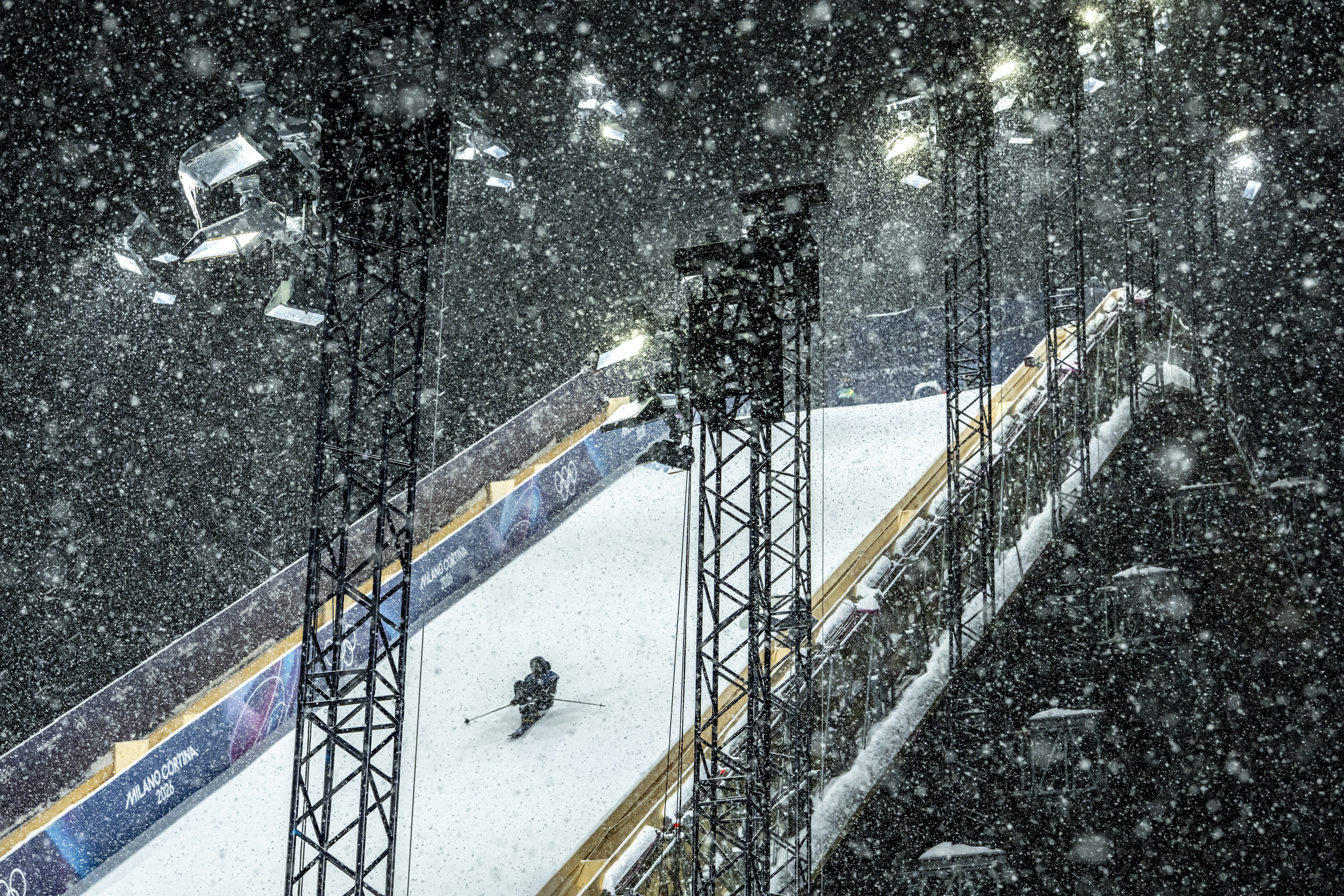 En skiløper raser ned en snødekt, opplyst bakke om natten, omgitt av høye flomlys og kraftig fallende snø