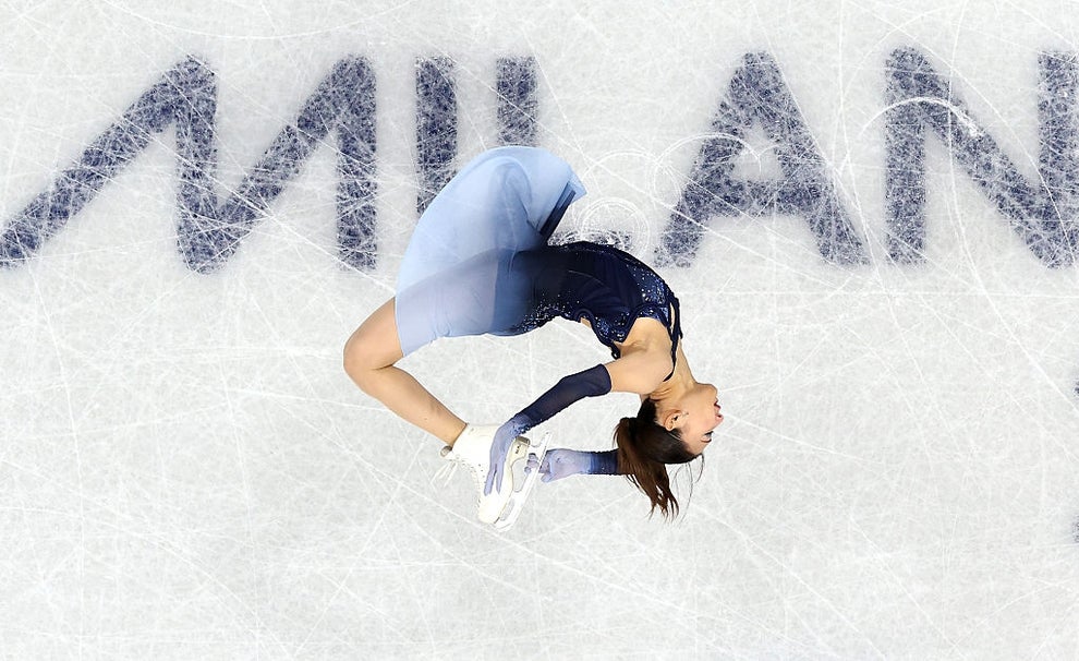 Figure skater mid-air, performing a backflip on ice, with the word "MILAN" visible beneath. The skater wears a stylish dress
