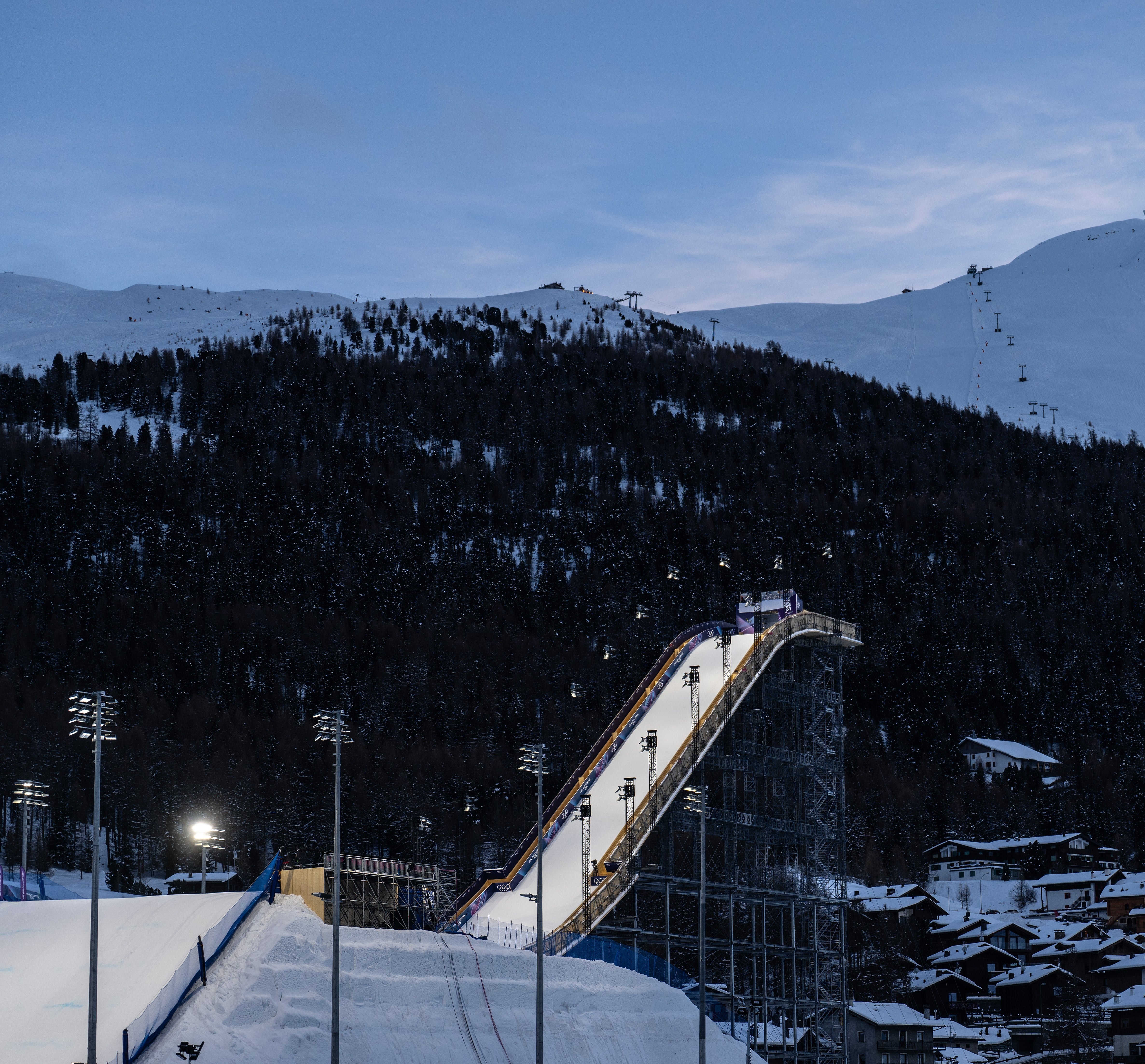 Hopprampe med skiløpere, omgitt av snødekte fjell og en landsby nedenfor, i skumringen