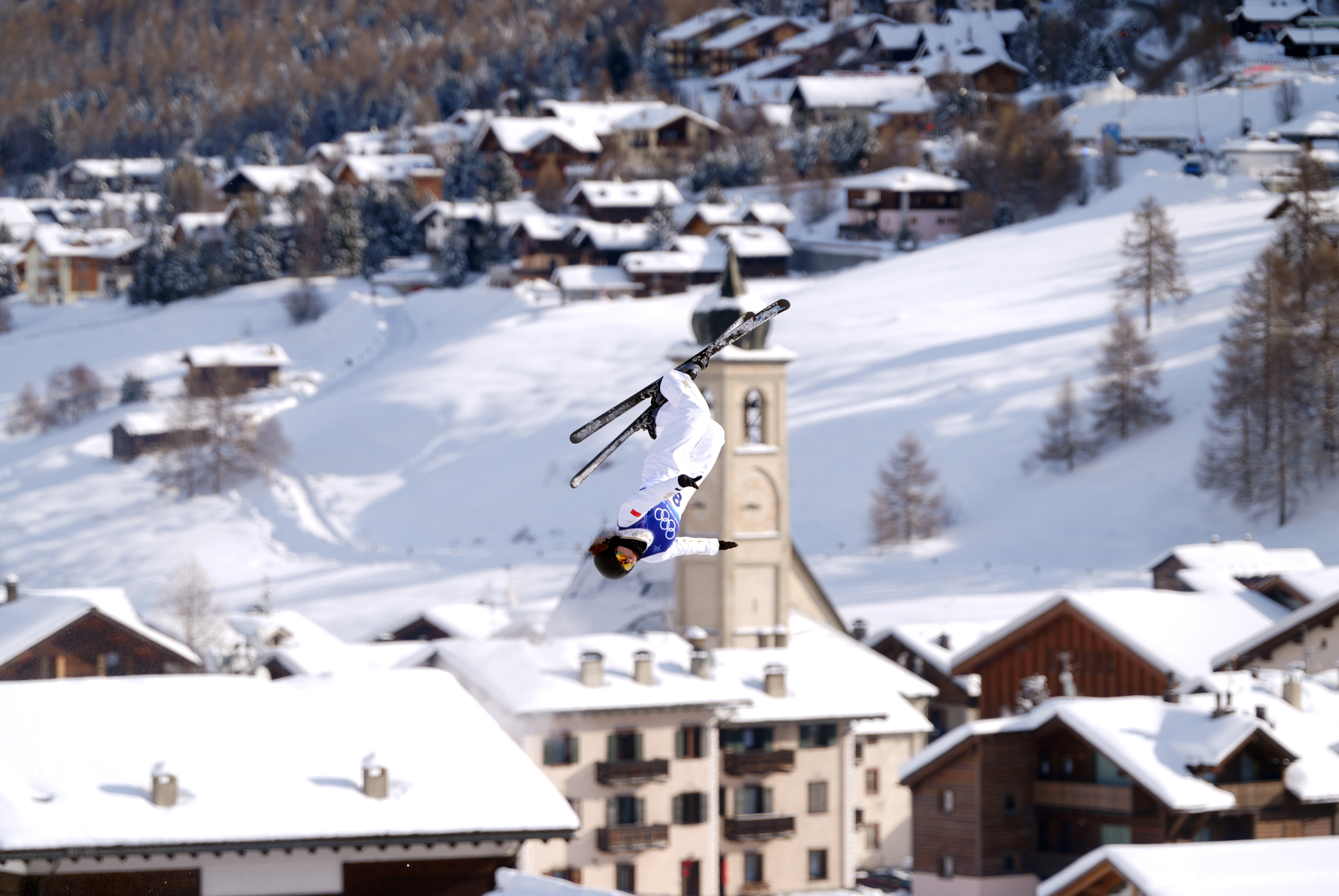 Skiløper utfører en backflip midt i luften over en snødekt alpinlandsby. Kirketår og hytter er synlige i bakgrunnen