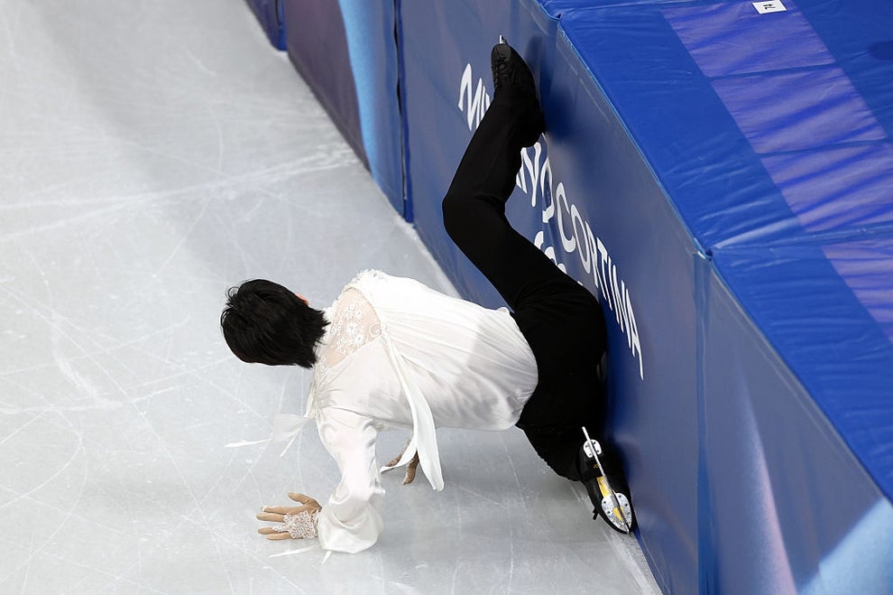 A figure skater in a white shirt has an awkward fall during a performance, sliding into the rink barrier with one leg propped up on it