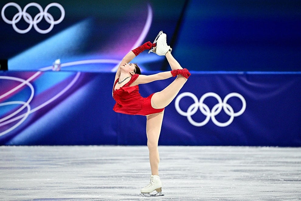 A figure skater performs a Biellmann spin on ice, wearing a stylish dress and matching accessories, with Olympic rings visible in the background