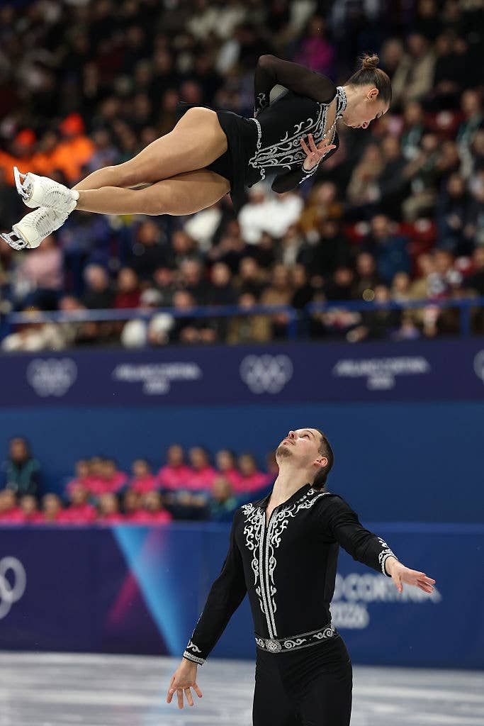 Figure skating duo performing a lift during a competition, with the female skater striking a pose mid-air, and both wearing intricately patterned outfits