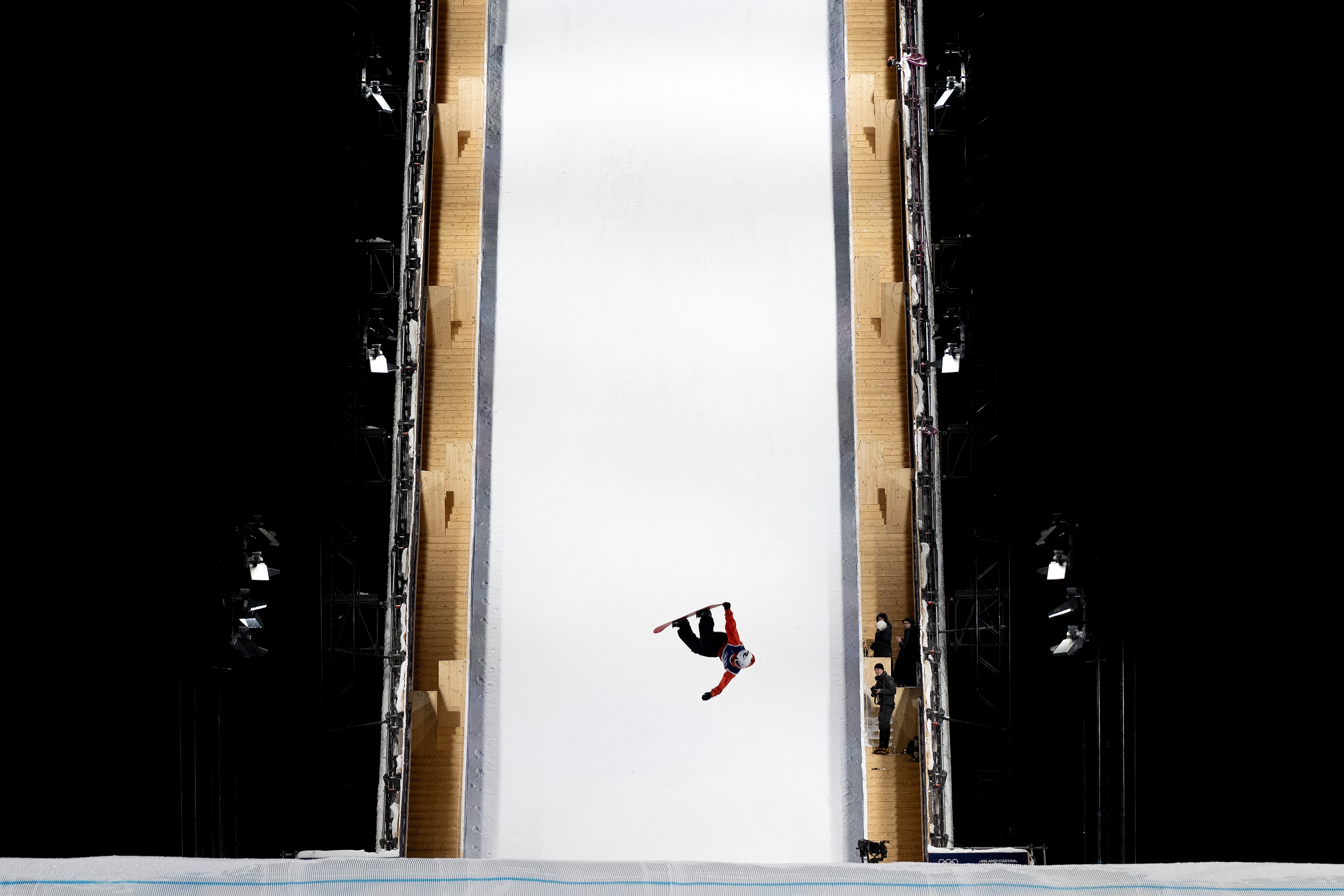 En snowboarder utfører en flip i luften på en stor rampe under en konkurranse, omgitt av tilskuere og lys