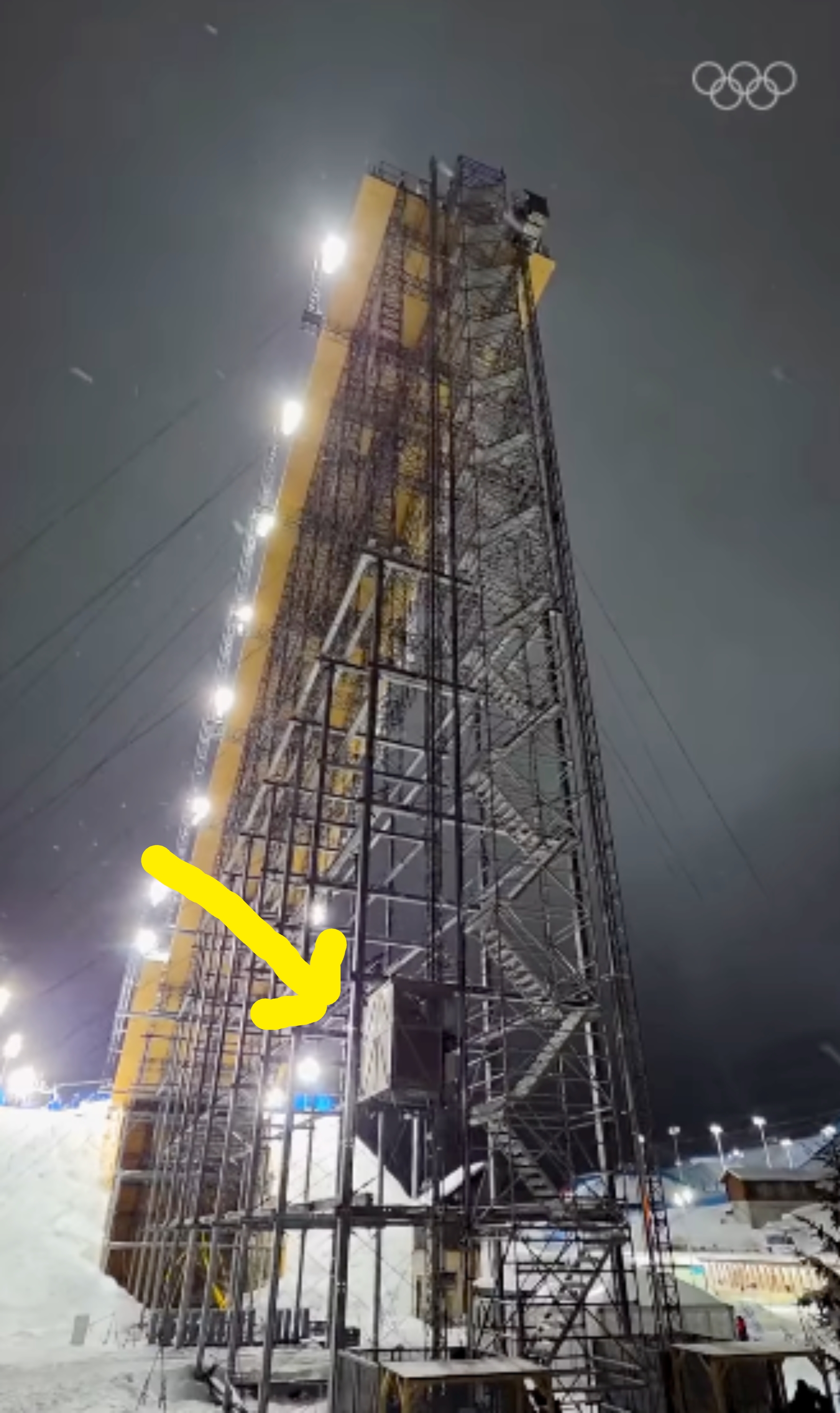 Høy hoppkonstruksjon ved et olympisk arrangement, opplyst av skarpe lys, med snødekte omgivelser og en mørk himmel i bakgrunnen