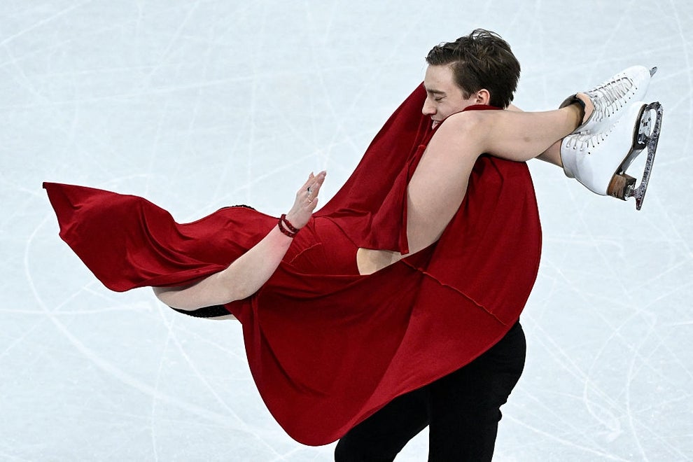 Two ice dancers perform a lift. The skater being lifted wears a flowing dress, creating dynamic movement in the performance