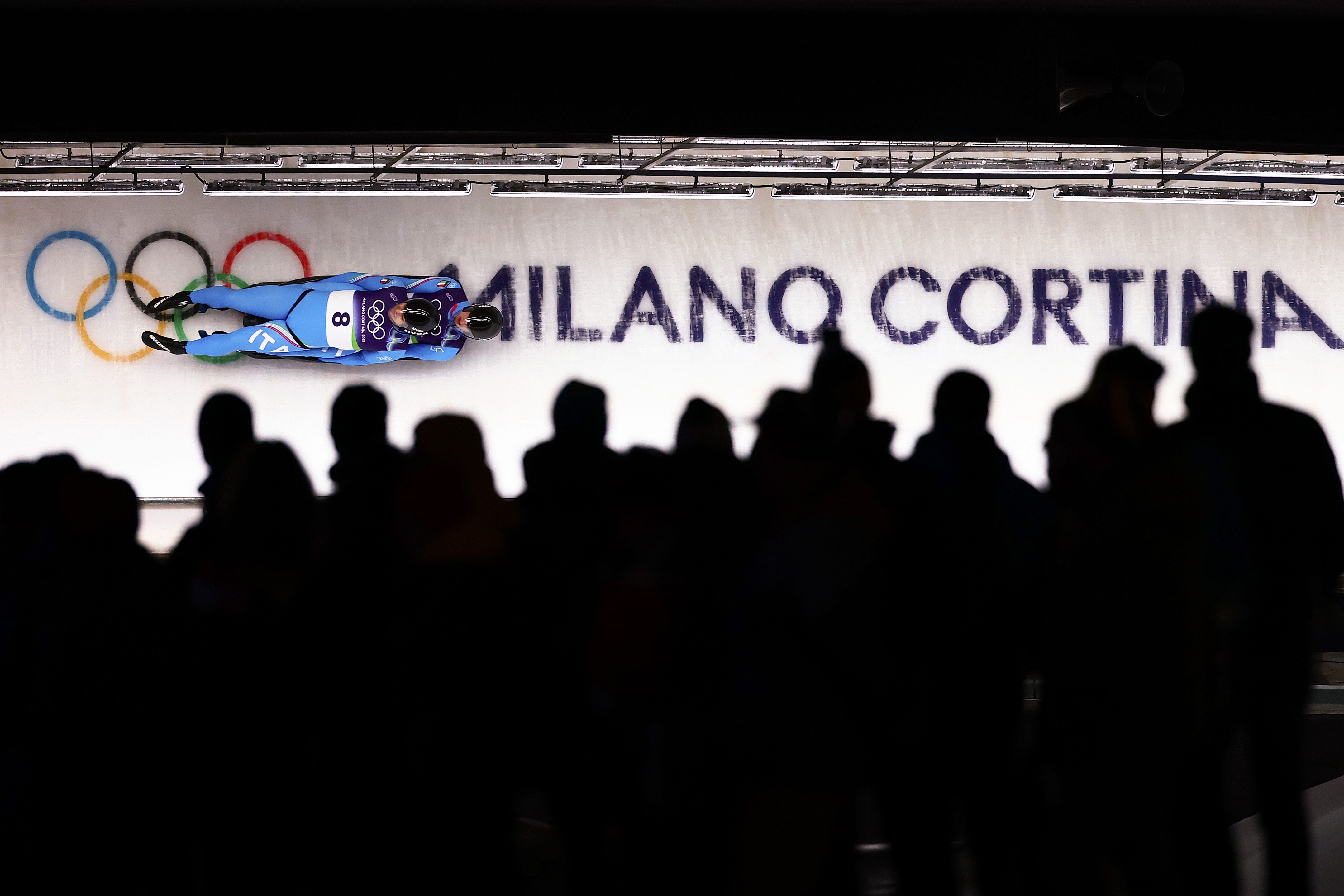 Idrettsutøver som konkurrerer i et akearrangement på Milano Cortina-arenaen med OL-ringer, tilskuere synlige i forgrunnen