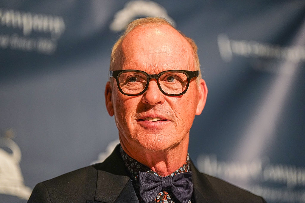 Person with glasses, wearing a suit   and bow tie, stands against a backdrop with logos