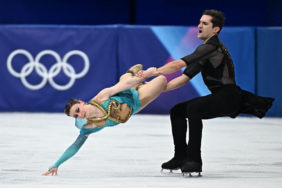 Figure skaters performing a dramatic lift on ice, with Olympic rings in the background. Skater in an embellished outfit; partner in a sleek, dark costume