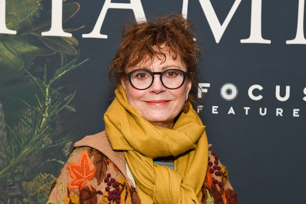 Person wearing glasses and a patterned overgarment   with a yellowish  scarf, lasting  successful  beforehand   of a backdrop with text