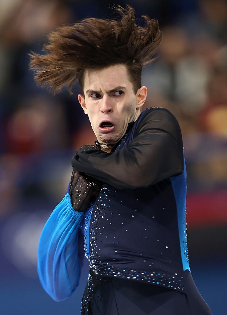Figure skater mid-performance, facial expression intense, wearing an embellished outfit with flowing sleeves. Hair captured in dramatic motion