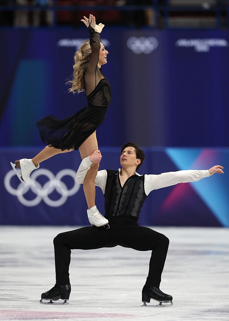Two figure skaters perform an intricate lift on the ice. The female skater wears a flowing outfit, and the male skater is in a fitted ensemble
