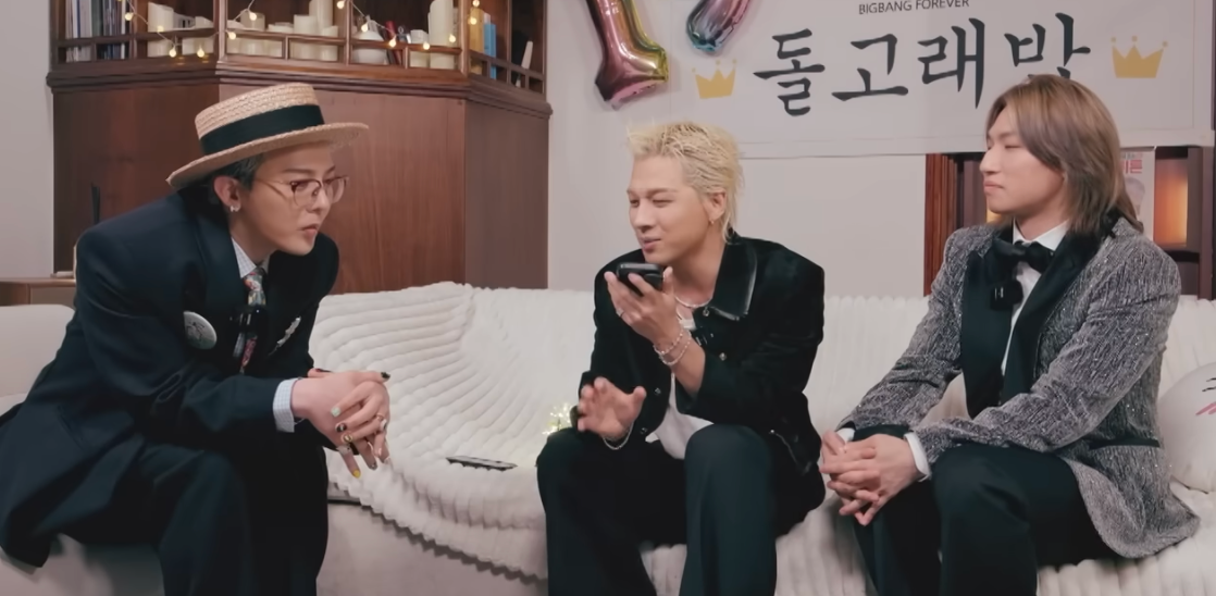 Tre musikere i dress sitter på en sofa i et musikkstudio, engasjert i samtale, mens en snakker i en telefon. Ballonger og et skilt dekorerer bakgrunnen