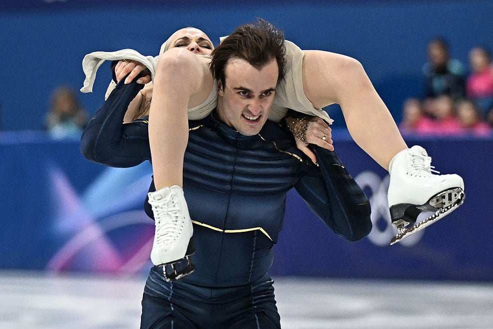 Figure skating pair performing a dynamic lift on the ice; the man is in a sleek outfit, carrying his partner on his shoulders during the routine