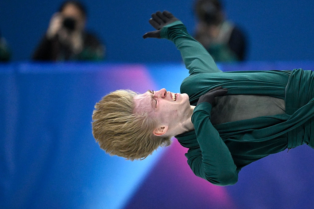 En kunstløper i et dynamisk, grasiøst spinn på isen, iført et elegant, flytende kostyme. Fotografer er synlige i bakgrunnen