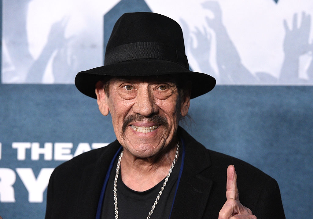 Person wearing a fedora and a necklace, smiling and pointing upward astatine  an lawsuit   with a patterned backdrop