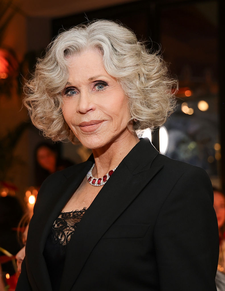 Person with wavy grey  hairsbreadth  successful  a achromatic  suit   and lace top, wearing a reddish  gemstone necklace, lasting  successful  an indoor lawsuit   setting