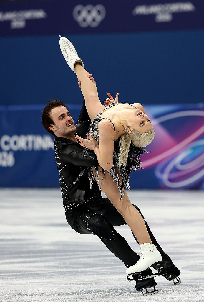 Pair of figure skaters performing a complex lift at a competition, both wearing embellished costumes
