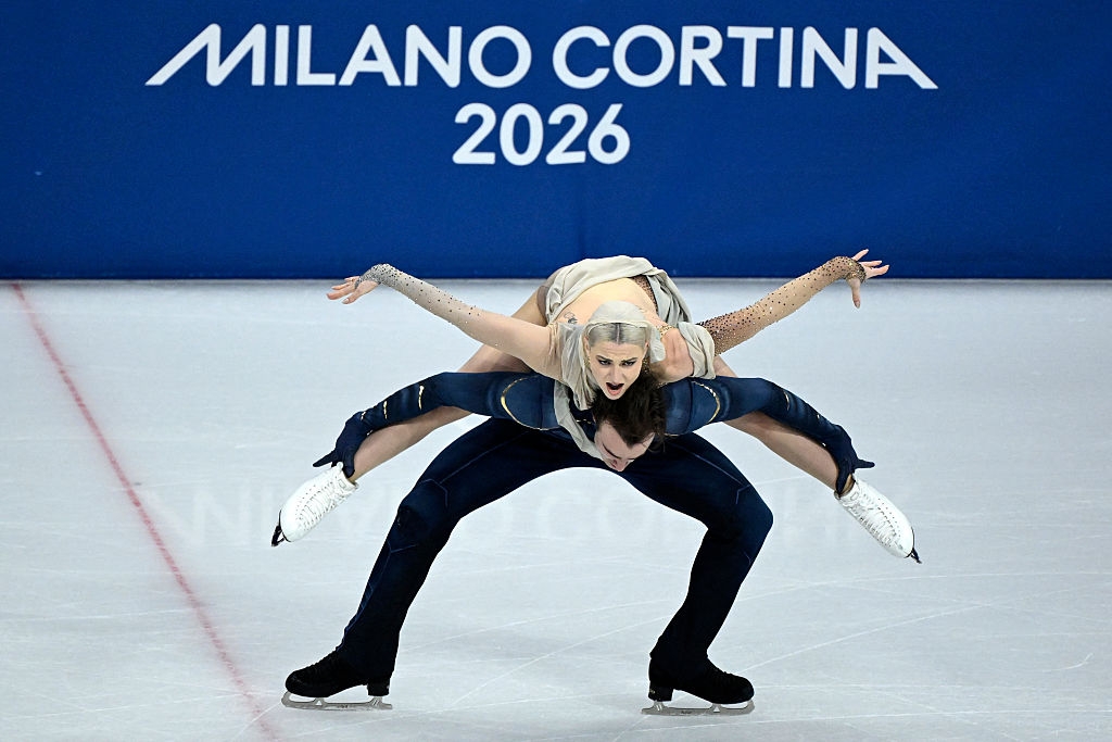 Kunstløpspar utfører et intrikat løft på Milano Cortina 2026-arrangementet, med skiltet synlig i bakgrunnen