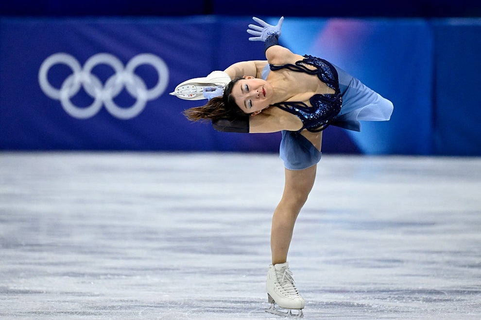 A figure skater performs a complex spin on the ice, showcasing athleticism and grace. Olympic rings are visible in the background