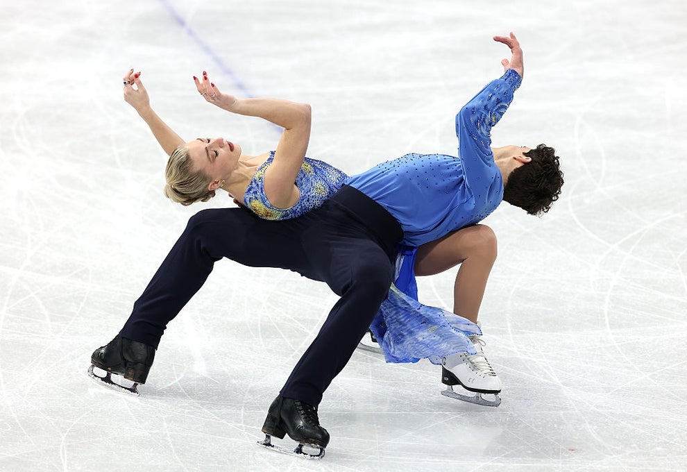 Two ice dancers mid-performance, one wearing a patterned top and dark pants, the other in a flowing dress with intricate detailing, striking a dynamic pose