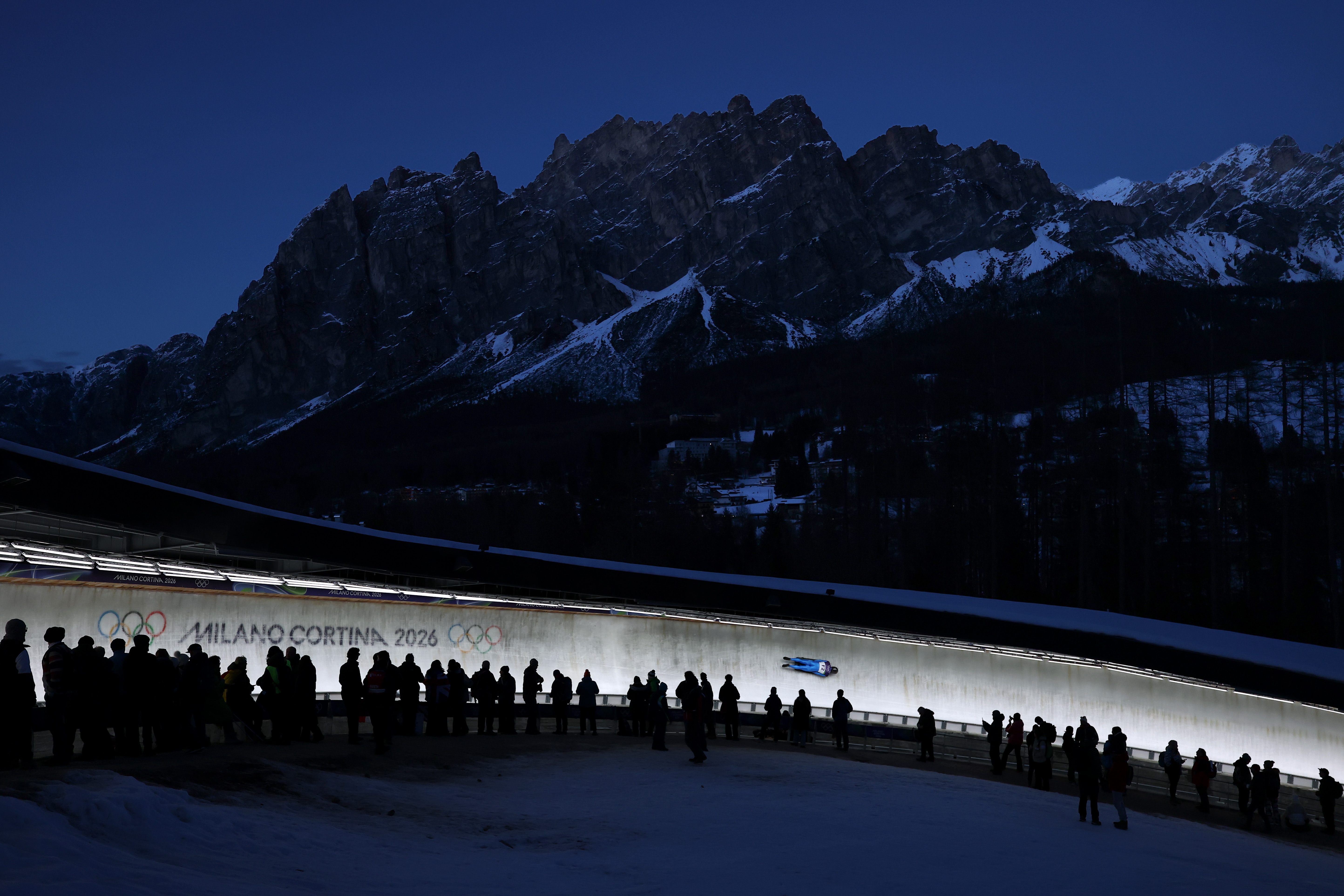 Tilskuere ser på en akeutøver som raser på en bane med fjell i bakgrunnen ved vinter-OL i Milano Cortina 2026