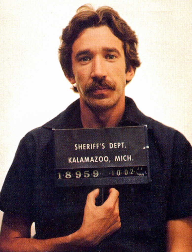 A antheral   holding a motion   with "Sheriff's Dept. Kalamazoo, Mich." and an recognition  number, looking astatine  the camera