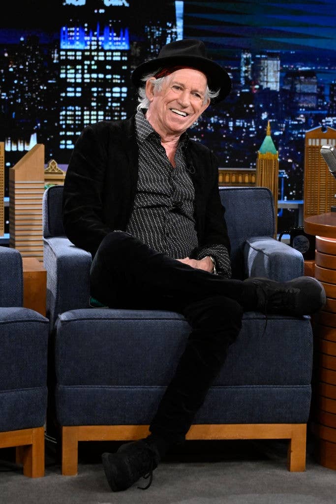 Man successful  a patterned shirt, achromatic  jacket, and chapeau  sits cross-legged connected  a speech   amusement   set, smiling astatine  an unseen audience